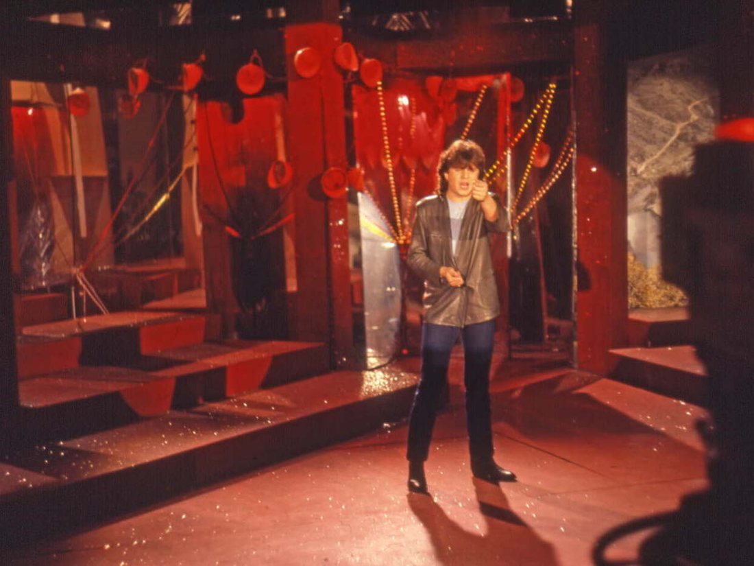 Daniel Balavoine, veste de cuir et micro à la main, chantant sur un plateau de télévision aux décors rouges