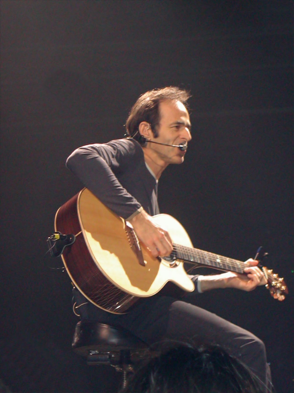 Jean-Jacques Goldman seated on a stool with an acoustic guitar and a headset microphone, in concert
